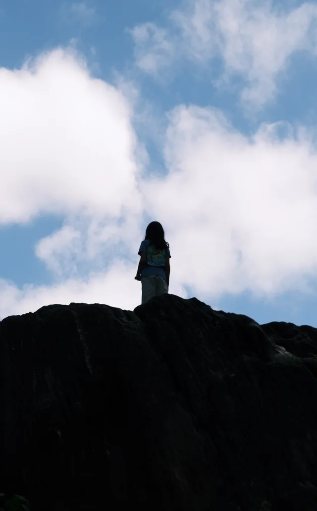 A small person stands on a dark ridge beneath bright clouds.