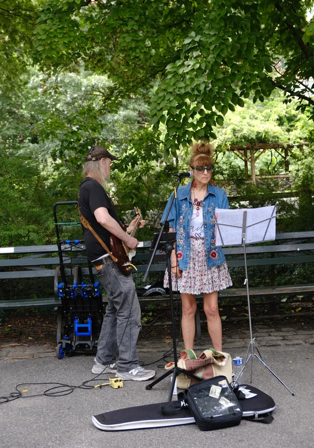 Two street musicians perform beside a music stand under a leafy canopy.