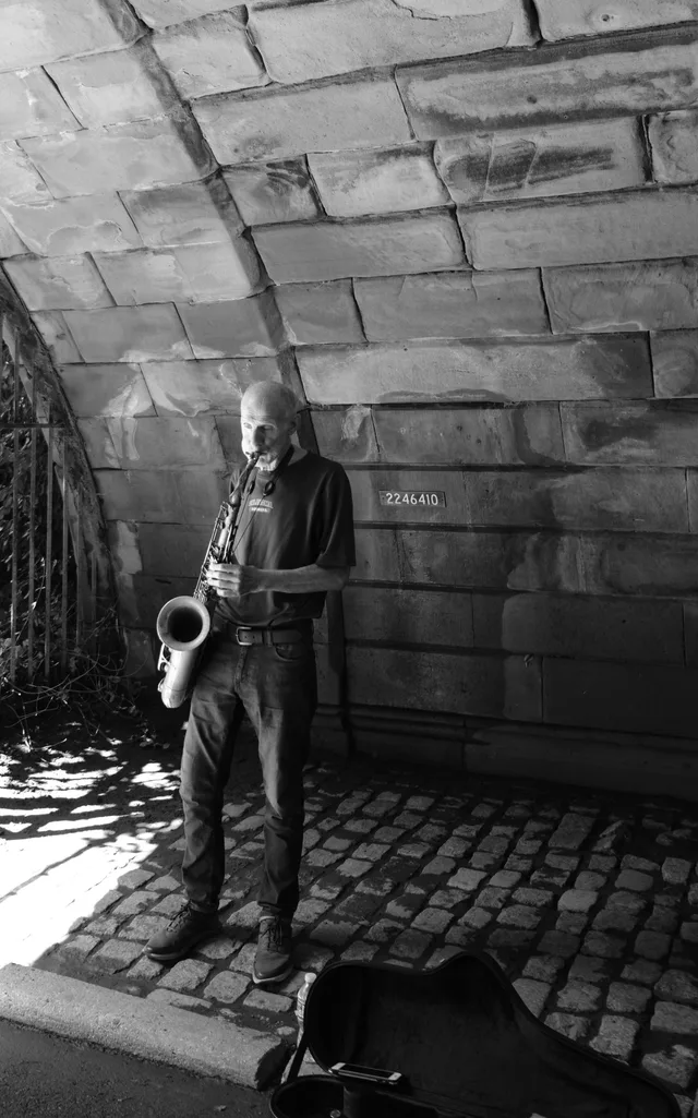 A saxophonist stands beneath a heavy stone arch in monochrome.