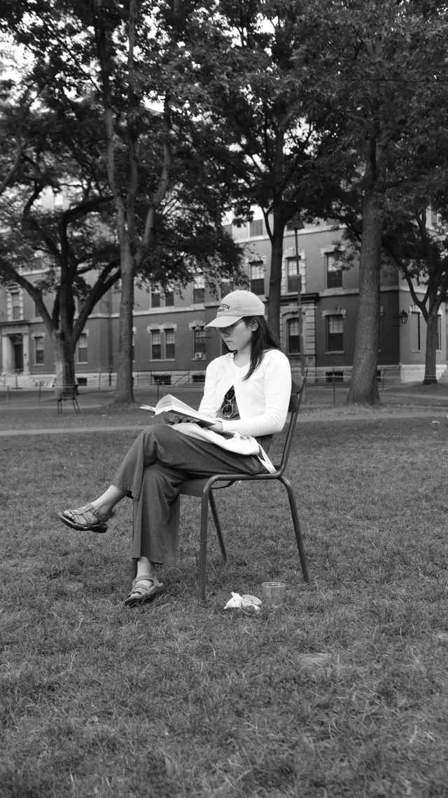 A hat-wearing reader sits alone on a lawn in monochrome.