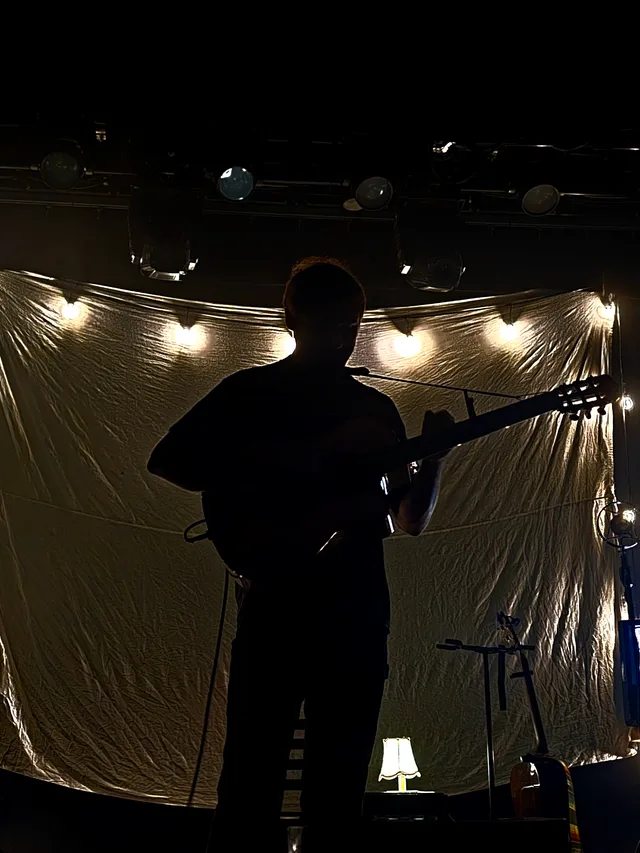 A guitarist appears almost entirely in silhouette before a lit backdrop.