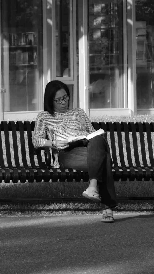 A reader sits alone on a slatted bench in monochrome.