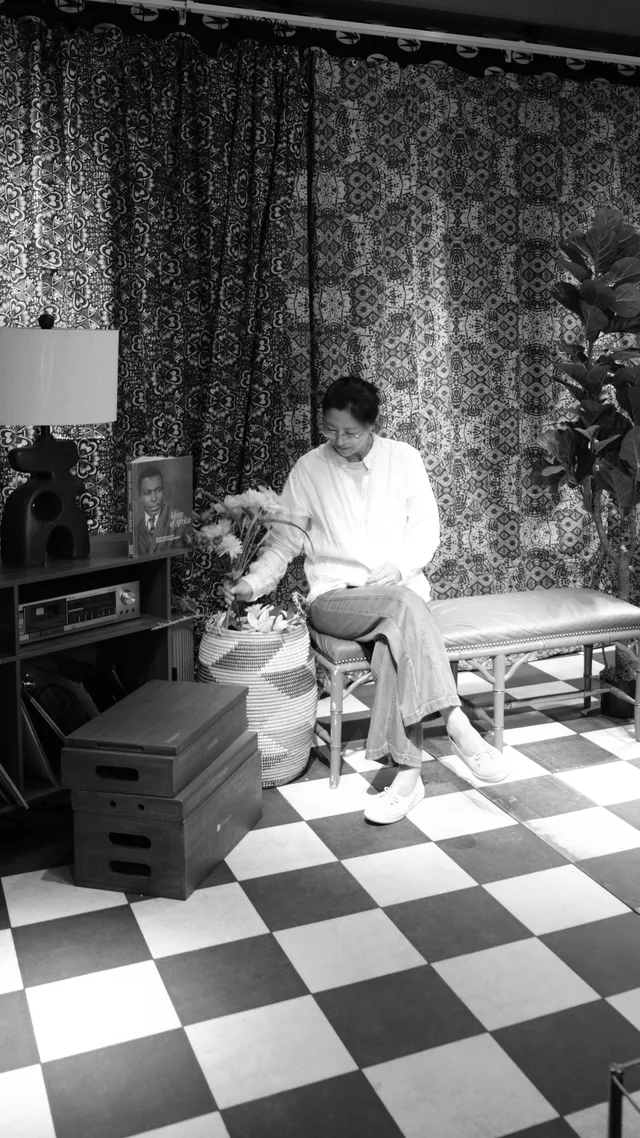 A seated figure reaches toward flowers beside a bench in a richly patterned room.