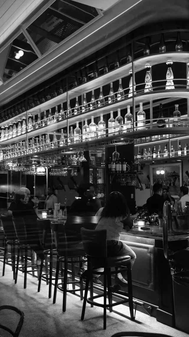 A monochrome bar is framed by long illuminated shelves and repeated stools.
